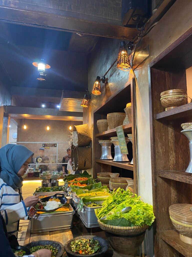 A person serving themselves at a buffet with various fresh vegetables and dishes in a rustic restaurant setting, featuring wicker basket decor and industrial-style hanging lights.