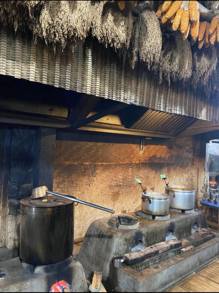 An indoor traditional kitchen with large pots on wood-fired stoves, bundles of dried corn hanging from the ceiling, and bamboo wall paneling in the background.