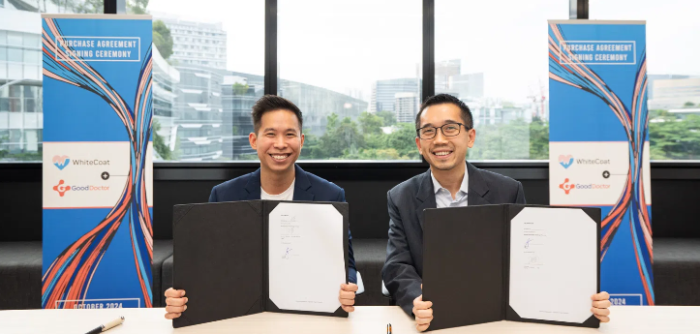 Two men smiling while holding signed agreements during a partnership signing ceremony, with backdrop banners featuring company logos.